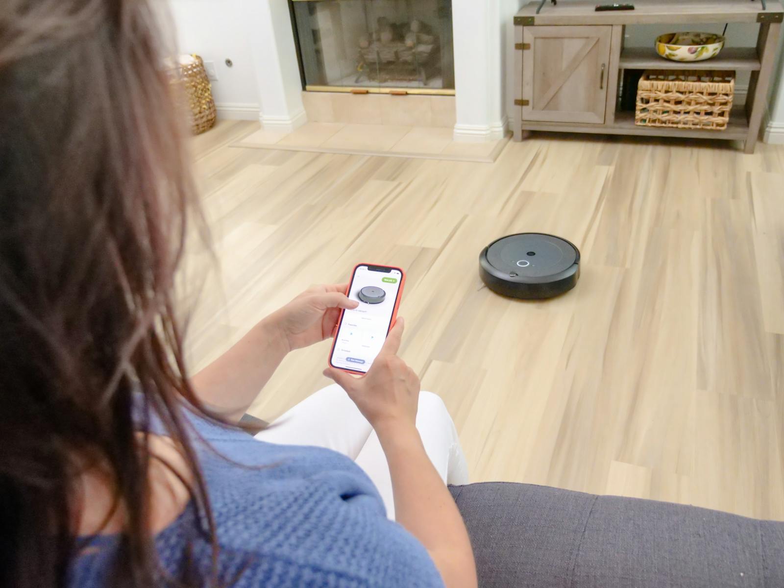 Overhead view of a woman controlling a robot vacuum using a smartphone app on a wooden floor.