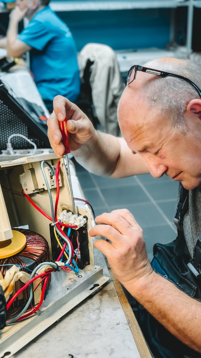 Senior electrician focused on fixing circuit board with red wires in a workshop setting.